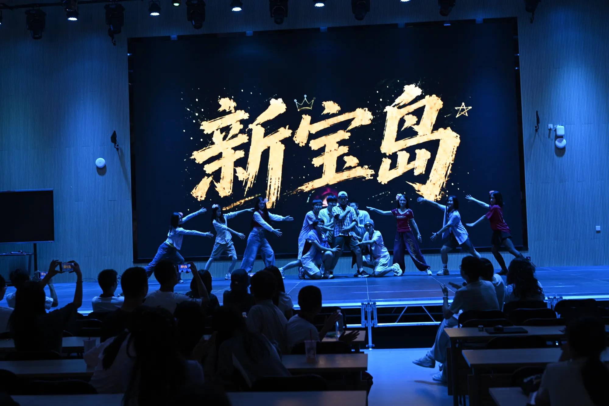 蓝色灯光照耀的舞台上，背景里是黄色新宝岛大字，舞台上一群人跳舞