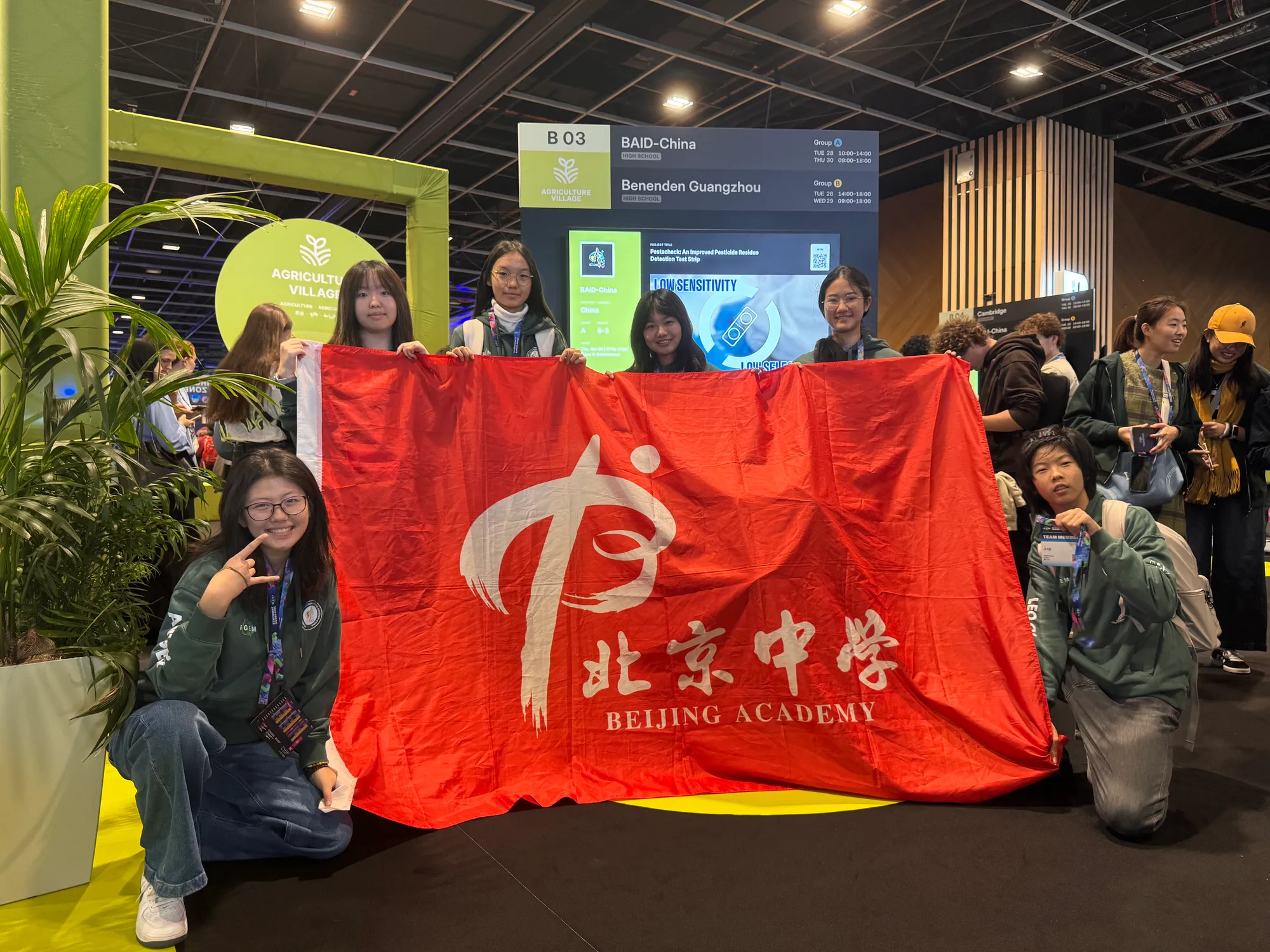 在 BAID-China 队伍的 iGEM 展板前,穿着队服的同学们举着北京中学旗帜合影